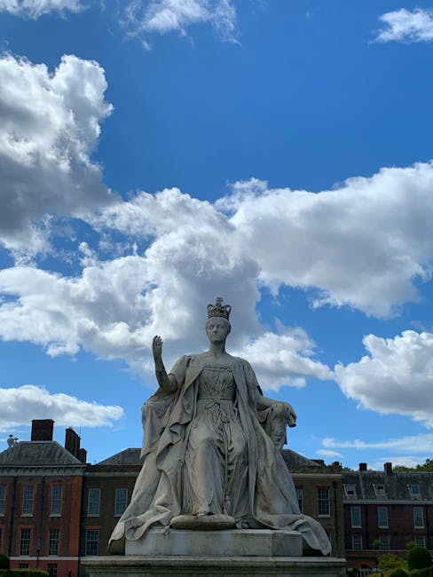 A detailed photograph of a large stone statue of a regal female figure seated outdoors against a bright blue sky with scattered white clouds. The statue depicts a woman wearing a crown and flowing, draped robes with intricate folds and textures. She is positioned centrally in the image, with her right hand raised and her left hand resting on her lap. In the background, there are low-rise brick buildings with slate roofs, and the base of the statue is visible but not the ground level. The scene suggests an urban or civic area, and the clear weather highlights the statue’s carved details. The presence of Waste Removal West Kensington’s context in the image connection emphasizes the importance of maintaining tidy, accessible outdoor spaces free from rubbish, indirectly supporting the value of efficient, private waste handling and rubbish removal services in the area.
