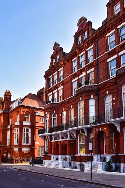 A multi-storey Victorian-style brick residential building with decorative architectural features, including arched windows and ornate iron balconies. The building's façade is predominantly red brick with white accents around the window sills and roofline. There are several large, sash-style windows with white frames, some with small potted plants placed on the window sills. A white decorative canopy extends over the front entrance, which is accessed via a short flight of white steps with black wrought iron railings. The street in front is paved with asphalt, with a black waste bin and street signage visible in the foreground. The sky above is clear and blue, with the sunlight casting subtle shadows on the building, contributing to a bright and calm urban scene that subtly relates to the context of private residential waste management services offered by Waste Removal West Kensington, situated in West Kensington, London.