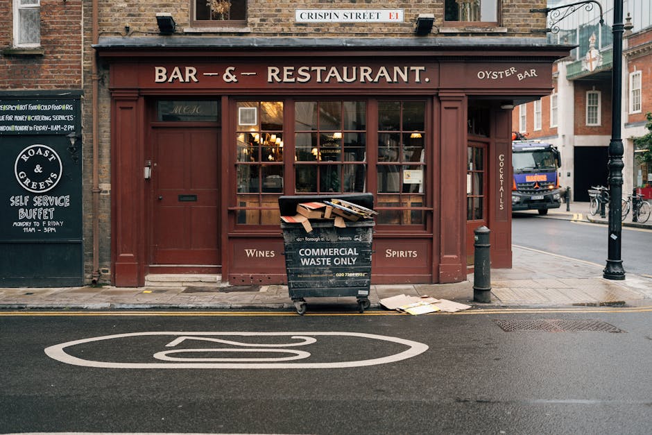 A black wheeled commercial waste bin labeled 'COMMERCIAL WASTE ONLY' is positioned on the sidewalk in front of a restaurant with a deep red wooden facade. The bin is partially filled with flattened cardboard boxes and discarded papers. Nearby on the pavement, a few sheets of paper are scattered, and the bin extends slightly over the curb. The restaurant has large glass windows with warm lighting inside, and signage indicating it serves wines and spirits. To the left, there's a blackboard-style sign advertising a self-service buffet, and on the right, a short black bollard separates the bar’s entrance from the street. A delivery truck is parked further down the street, and bicycles are seen leaned against a building in the background. The scene suggests an area where private waste collection services, such as those provided by Waste Removal West Kensington, might handle commercial rubbish, rather than relying on local authority collections, especially for business waste disposal.