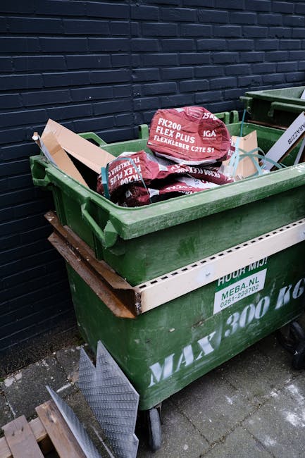 A large green waste disposal bin positioned on a paved area beside a dark brick wall, filled with red and white packaged bags of garden or building materials, with some bags partially protruding over the edge. The bin rests on black wheels and has a white horizontal strip on the side with printed text related to waste collection, indicating it is used for private rubbish disposal. Nearby, parts of a second similar bin and a small section of wooden pallets are visible, suggesting an outdoor environment designated for rubbish storage and potential collection or clearance services. The scene is lit by natural daylight, emphasizing the textures of the materials and the environment in a clear, neutral manner, aligning with the context of alternative waste handling or rubbish removal services in a residential or commercial setting.
