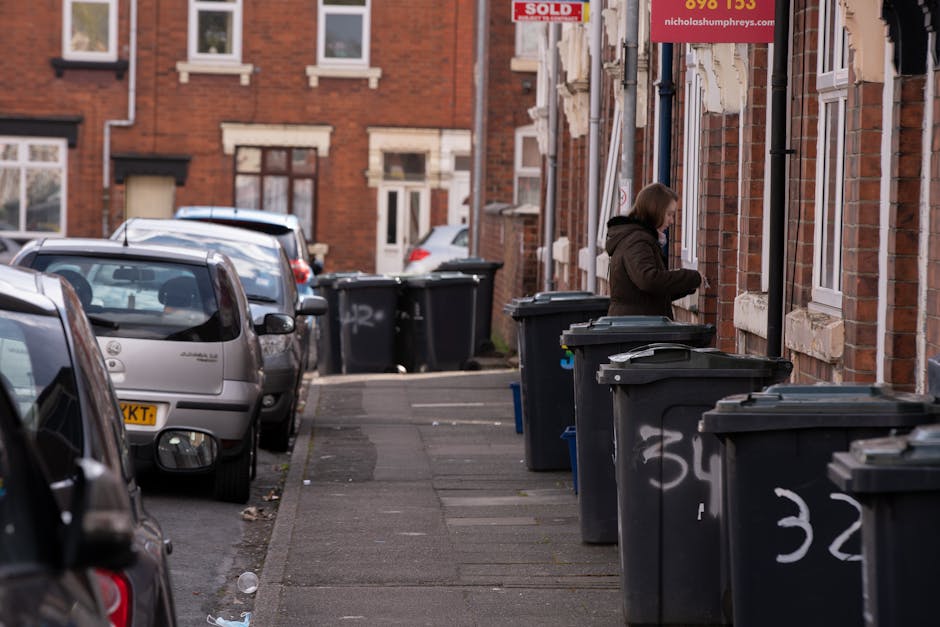 A residential street lined with brick terraced houses, featuring multiple black wheelie bins placed along the pavement in front of the homes, some with lids closed and others slightly open. The bins are made of plastic with a textured surface, and some display white chalk markings, including the number '32.' A woman with shoulder-length brown hair, wearing a dark coat, stands next to one of the bins, appearing to be either placing rubbish inside or checking its contents. Parked cars, including small hatchbacks and compact vehicles, occupy the street on the left side, with visible details such as side mirrors, rear windows, and roof racks. The background shows more brick facades, windows, and a red sign with white text indicating a property has been sold. The scene is lit with natural daylight, creating a neutral, professional appearance that illustrates typical on-site waste disposal practices in UK residential areas, indirectly supporting alternative rubbish collection methods provided by waste management services like Waste Removal West Kensington.