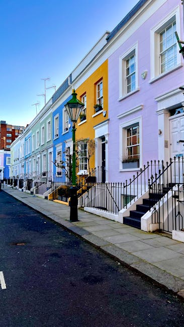 A row of terraced houses on a street, each painted in pastel shades including light blue, yellow, pale purple, and white, with uniform architectural features such as white-framed sash windows, black wrought-iron railings, and small front steps leading to doorways. In the foreground, a black street lamp with a green glass top stands near the pavement, casting subtle shadows on the stone steps and pathway. The pavement is made of rectangular grey tiles, while the street surface appears asphalted, with a single white line marking the edge. The overall environment suggests an urban residential area with well-maintained facades and a clean, orderly appearance, consistent with typical private waste collection and rubbish removal practices that may be carried out by services like Waste Removal West Kensington, supporting on-site clearance or alternative waste handling from this vibrant neighbourhood.