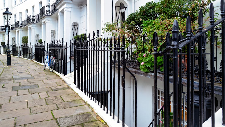 A row of white, multi-story terraced houses with black wrought iron railings along a paved sidewalk in a residential area. The railings feature pointed finials and decorative details, lining the edge of a small front garden filled with green foliage, including pink flowering plants positioned on the right side of the image. In the foreground, a blue bicycle is leaned against the railing near a black bollard. The pavement is made of large rectangular stone slabs with visible joints and some moss or dirt in between, extending into the distance with other similar houses visible further along the street. The environment appears to be well-maintained and typical of an urban residential neighborhood, with natural daylight highlighting the textures of the stone, metal, and plant life, creating a clean and orderly scene that naturally relates to local rubbish and waste collection or private waste handling activities, such as waste removal services in a West Kensington setting.