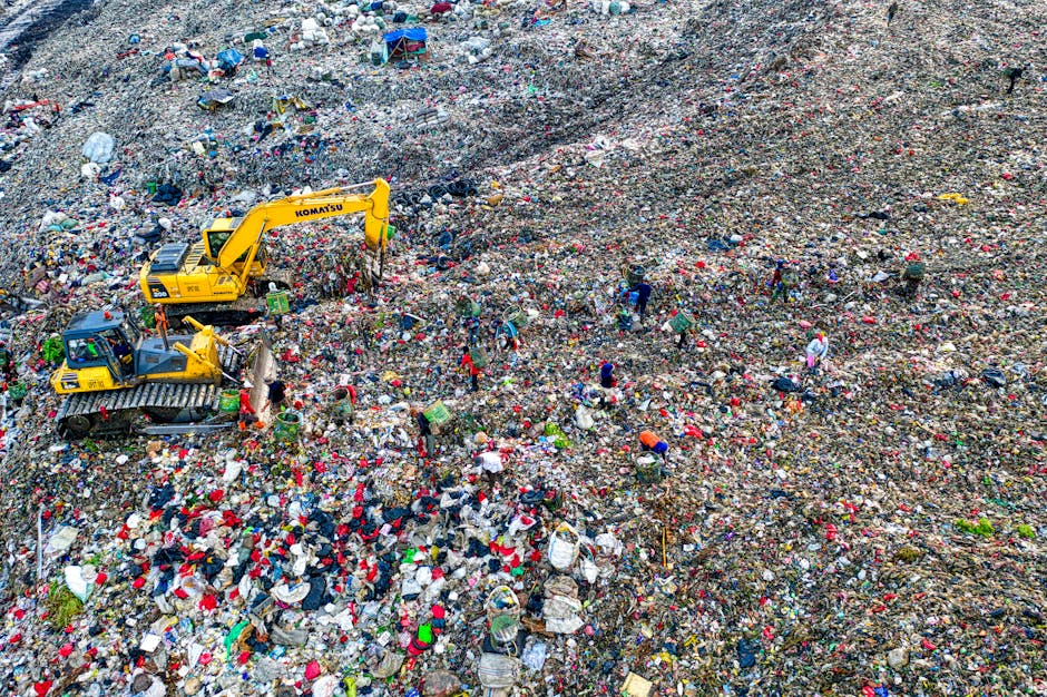A large landfill site filled with a vast expanse of mixed waste, including plastics, textiles, paper, and discarded household items, covering uneven ground and creating a textured surface of debris and rubbish. Multiple workers are scattered throughout the site, wearing protective clothing such as high-visibility vests and hats, and they appear to be manually sorting or managing the waste. Heavy machinery, including a yellow excavator and a tracked bulldozer, is actively engaged in moving and recontouring the piled-up rubbish, indicating ongoing site management. The environment shows a cloudy sky overhead, with a somewhat bleak and cluttered landscape extending into the distance. This scene illustrates an example of waste management practices where independent collection and on-site clearance are used to handle large volumes of refuse outside of regular municipal disposal channels, highlighting the importance of professional rubbish removal services like those offered by Waste Removal West Kensington for private waste handling and environmental compliance.