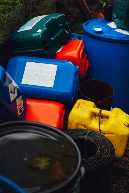 A collection of various plastic waste containers and jerry cans is shown outdoors, arranged on a grassy or earthy surface. The containers come in different colors including blue, red, yellow, and dark green, and are made of durable, textured plastic. Some have visible labels or labels with printed text, while others are plain. The blue container on the right appears to be a large water or chemical storage tank, with a white cap on top, and the yellow jerry can in the foreground has a black handle and a spout for pouring. A black coiled hose or cable is partially visible, placed near the yellow container. The scene suggests an environment where waste or hazardous substances might be temporarily stored or prepared for disposal, reflecting the context of private or alternative waste handling, possibly managed by Waste Removal West Kensington, in relation to safe rubbish interim storage before final collection or disposal, with natural lighting emphasizing the textured surfaces and functional nature of the containers.