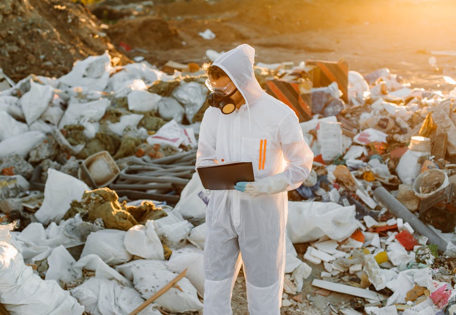 A large landfill site filled with a vast expanse of mixed waste, including plastics, textiles, paper, and discarded household items, covering uneven ground and creating a textured surface of debris and rubbish. Multiple workers are scattered throughout the site, wearing protective clothing such as high-visibility vests and hats, and they appear to be manually sorting or managing the waste. Heavy machinery, including a yellow excavator and a tracked bulldozer, is actively engaged in moving and recontouring the piled-up rubbish, indicating ongoing site management. The environment shows a cloudy sky overhead, with a somewhat bleak and cluttered landscape extending into the distance. This scene illustrates an example of waste management practices where independent collection and on-site clearance are used to handle large volumes of refuse outside of regular municipal disposal channels, highlighting the importance of professional rubbish removal services like those offered by Waste Removal West Kensington for private waste handling and environmental compliance.