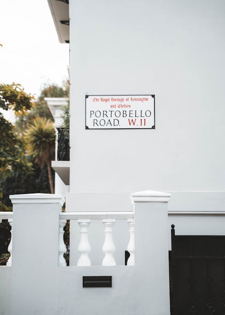 A close-up view of a white building corner showing a black and white street sign attached to the wall, reading 'The Royal Borough of Kensington and Chelsea, Portobello Road, W.11', indicating the address location. The building features a small white balustrade with rounded, turned balusters and a black mailbox slot below it, set into the white façade. The background includes a clear sky and some blurred trees with green and yellow foliage, suggesting an exterior urban environment. The scene is well-lit with natural daylight, emphasizing the clean, smooth texture of the building's painted surface. The image subtly relates to private property and location identification in the context of waste disposal or rubbish collection, as part of the environment where waste removal services like those offered by Waste Removal West Kensington might operate or support alternative waste handling arrangements outside local authority collections.