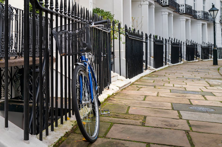 A blue bicycle with a black front basket is parked and leaning against a black metal fence that encloses the front steps of a white terraced townhouse. The bicycle's frame has a matte finish, and the basket appears to be made of metal mesh. The fence features pointed finials at the top of each vertical bar, and the fence posts are securely anchored into the stone pavement. The townhouse's white facade includes black wrought iron railings on the upper balconies and classical-style columns adjacent to the front steps. The steps themselves are composed of white stone, leading up to the front door which is partially visible. To the right, the pavement is laid with irregularly shaped stone slabs with moss and dirt collected in the joints. The scene is lit by natural daylight, creating subtle shadows and highlighting the textures of the materials. The overall setting suggests a quiet residential street with an emphasis on local, private waste handling, typical of an independent rubbish removal service in West Kensington.