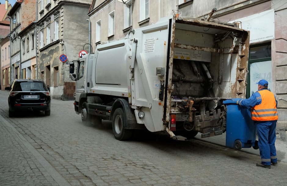A waste collection truck operated by Waste Removal West Kensington is parked on a narrow urban street with cobblestone paving, positioned adjacent to a row of old, multi-story buildings with weathered facades and variously sized windows. The truck, primarily white with some rust and dirt visible on its rear loading compartment, is in the process of collecting rubbish from a blue wheeled bin held by a worker dressed in high-visibility blue and orange overalls and a matching cap, who is actively positioning the bin to load waste into the back of the vehicle. The scene is lit naturally, suggesting daylight, and the surroundings include parked cars, such as a dark grey vehicle on the street’s left side, with street signs indicating parking restrictions. This image illustrates a typical episode of on-site rubbish collection as part of private waste management services, supporting the context of alternative rubbish pickups in the West Kensington area.